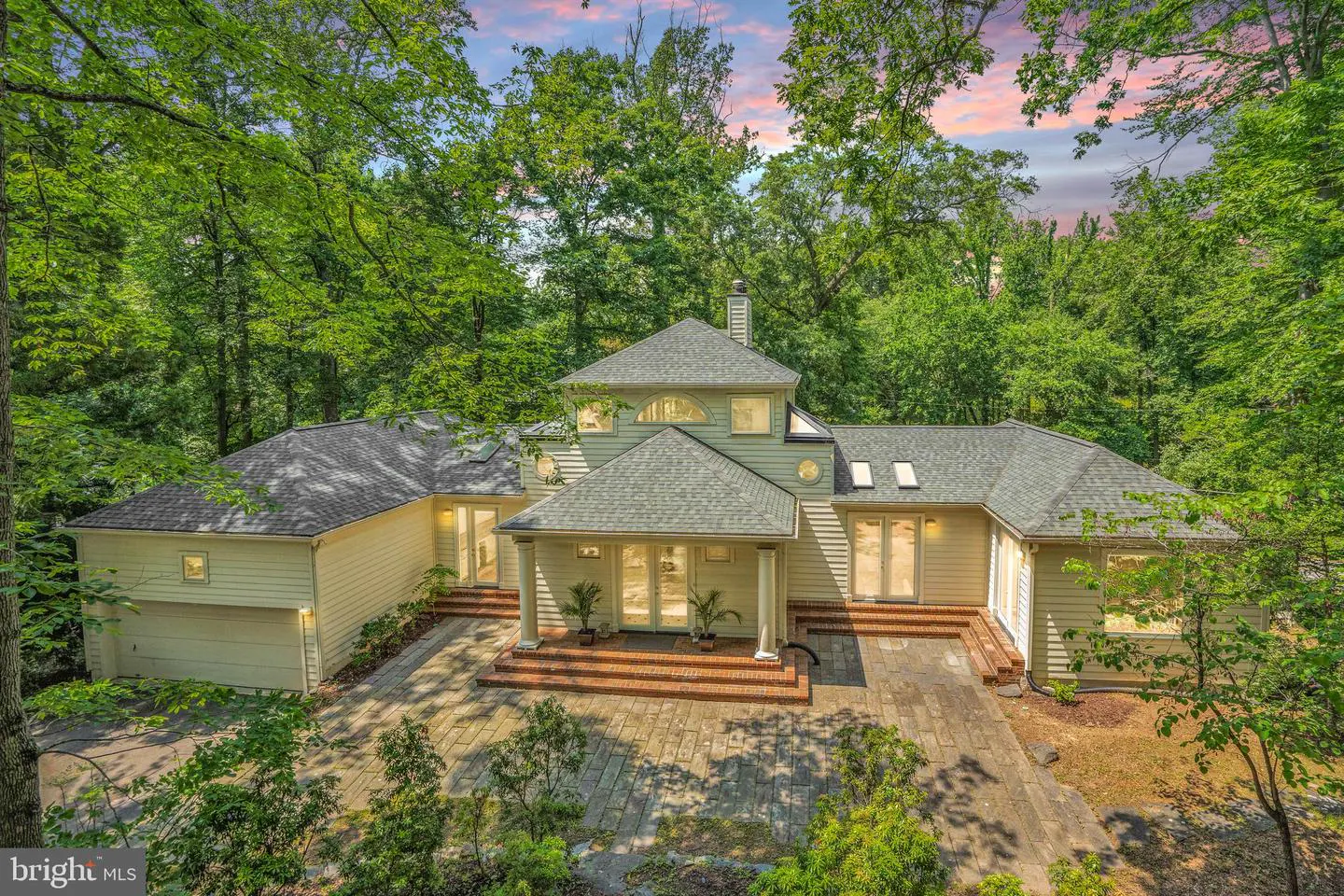Aerial view of a beige house with a gray roof, surrounded by green trees and a stone patio.