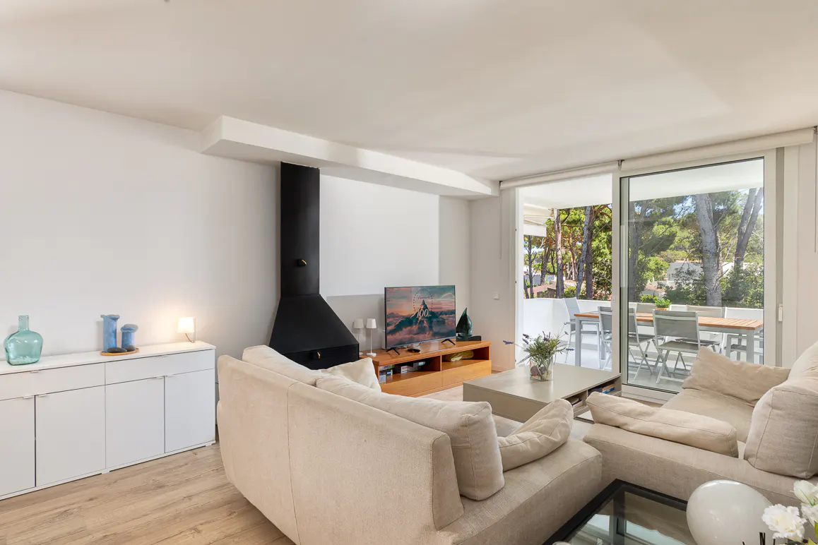Modern living room with beige sofas, a black fireplace, TV on a wooden stand, and a sliding door to a patio with trees.