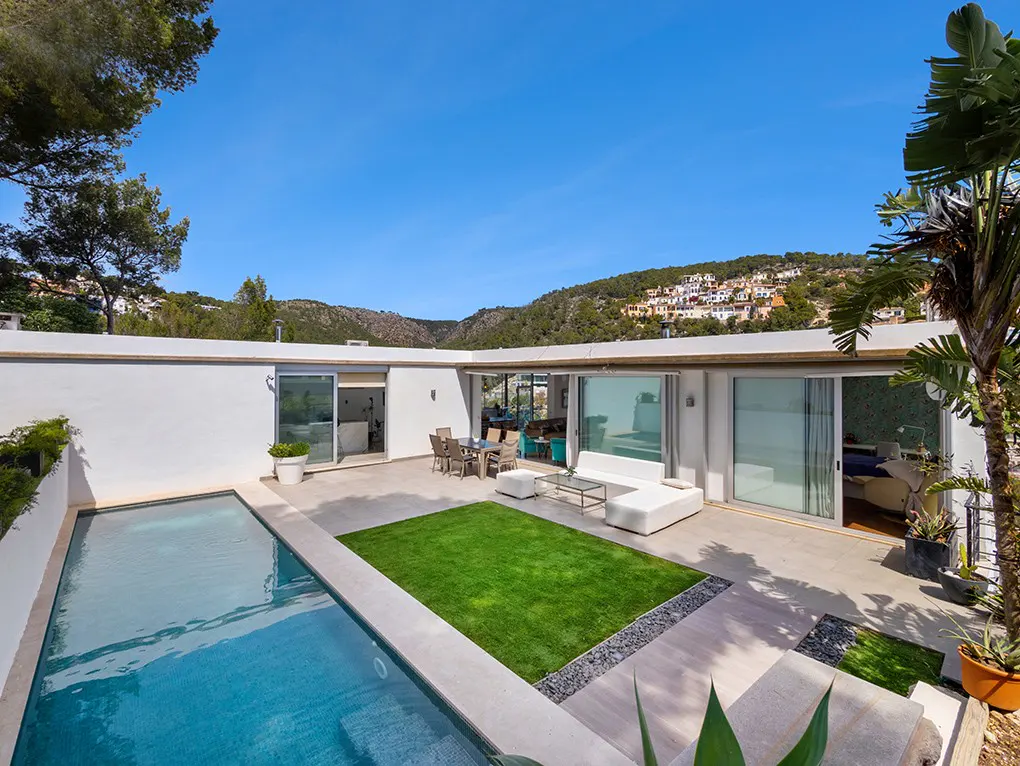 Modern villa patio with a swimming pool, green lawn, outdoor dining set, and white sofa against a hillside backdrop.