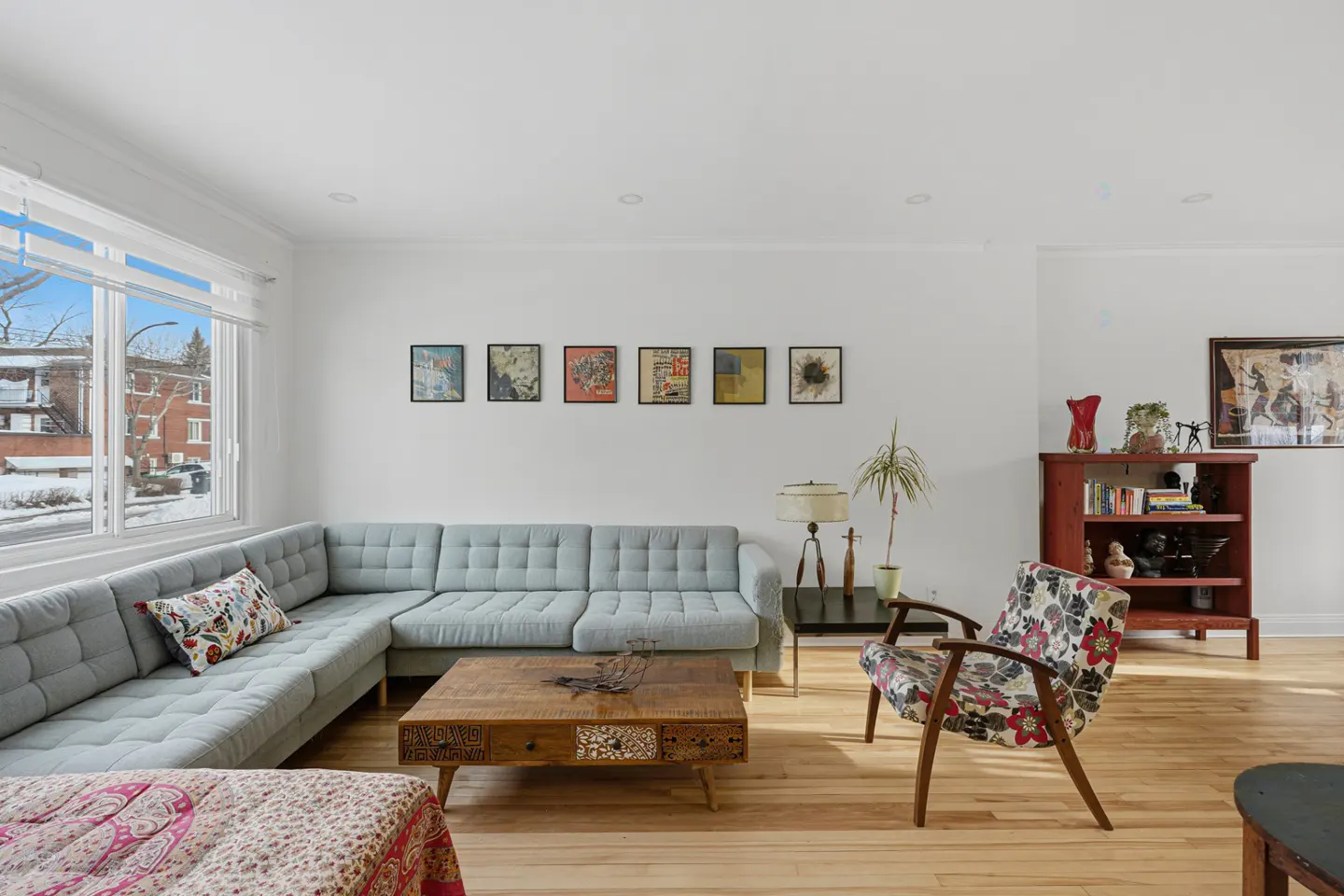 Bright living room with a light blue sectional sofa, wooden coffee table, floral armchair, and a red bookcase against a white wall.