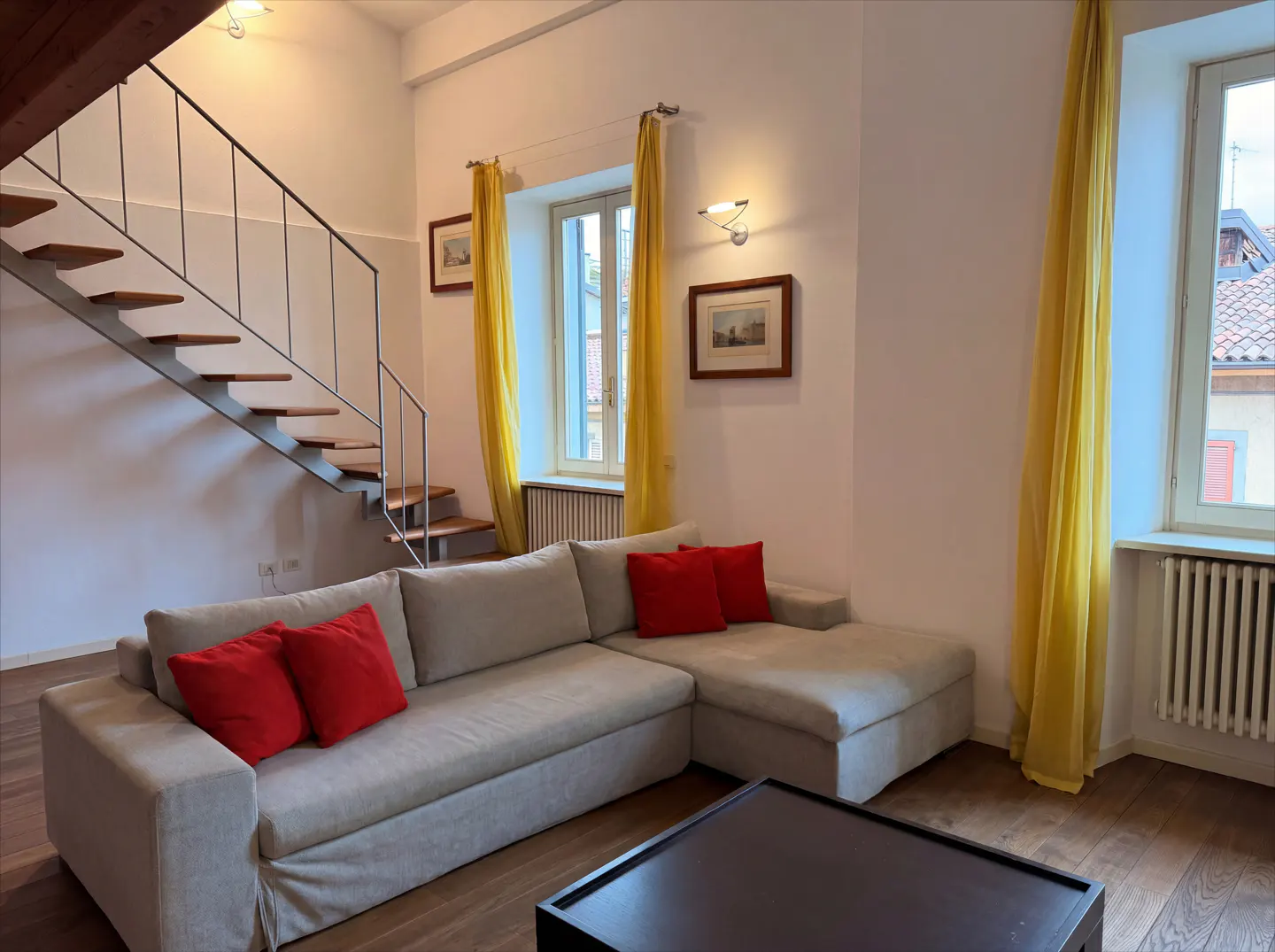 Living room with a beige sectional sofa with red pillows, a dark coffee table, and a staircase with wooden steps. Yellow curtains frame the windows.