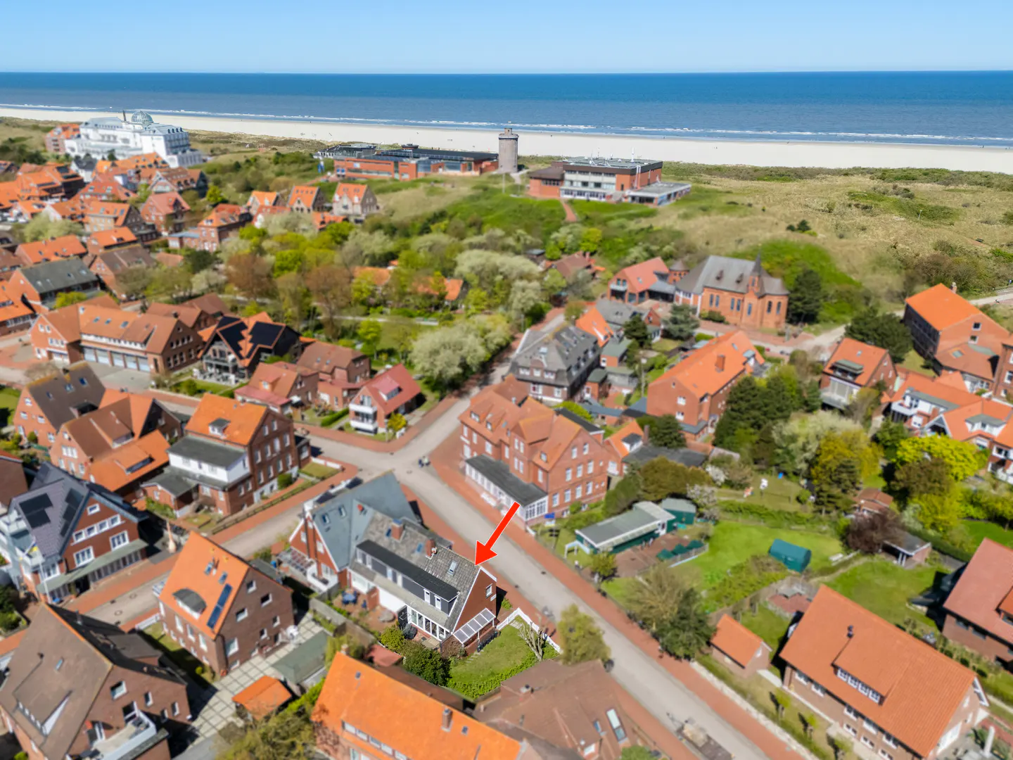 Aerial view of a coastal town with red-roofed buildings, an arrow points to a house with solar panels, and the ocean is in the background.
