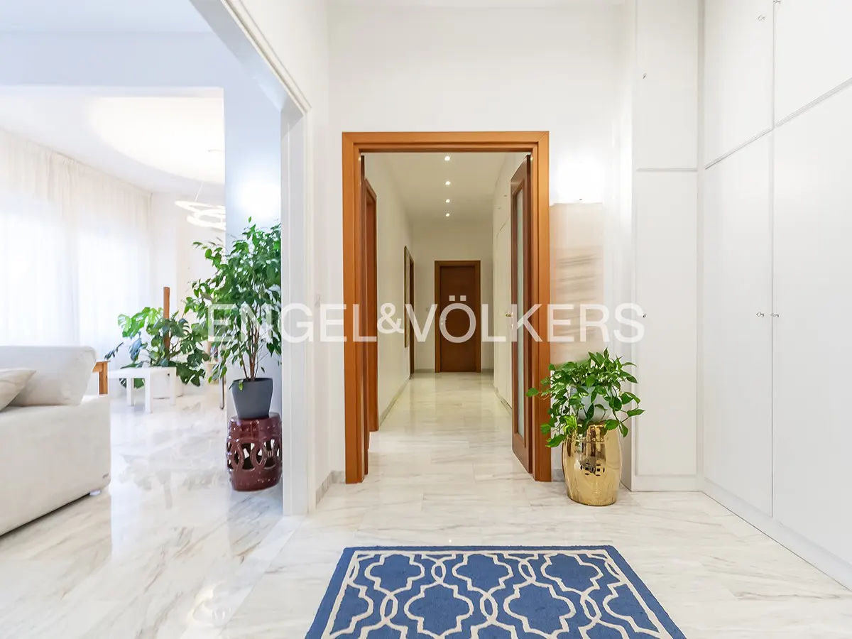 Bright hallway with marble floors, wood doors, and white walls. A blue patterned rug sits at the entrance. Plants add a touch of green.