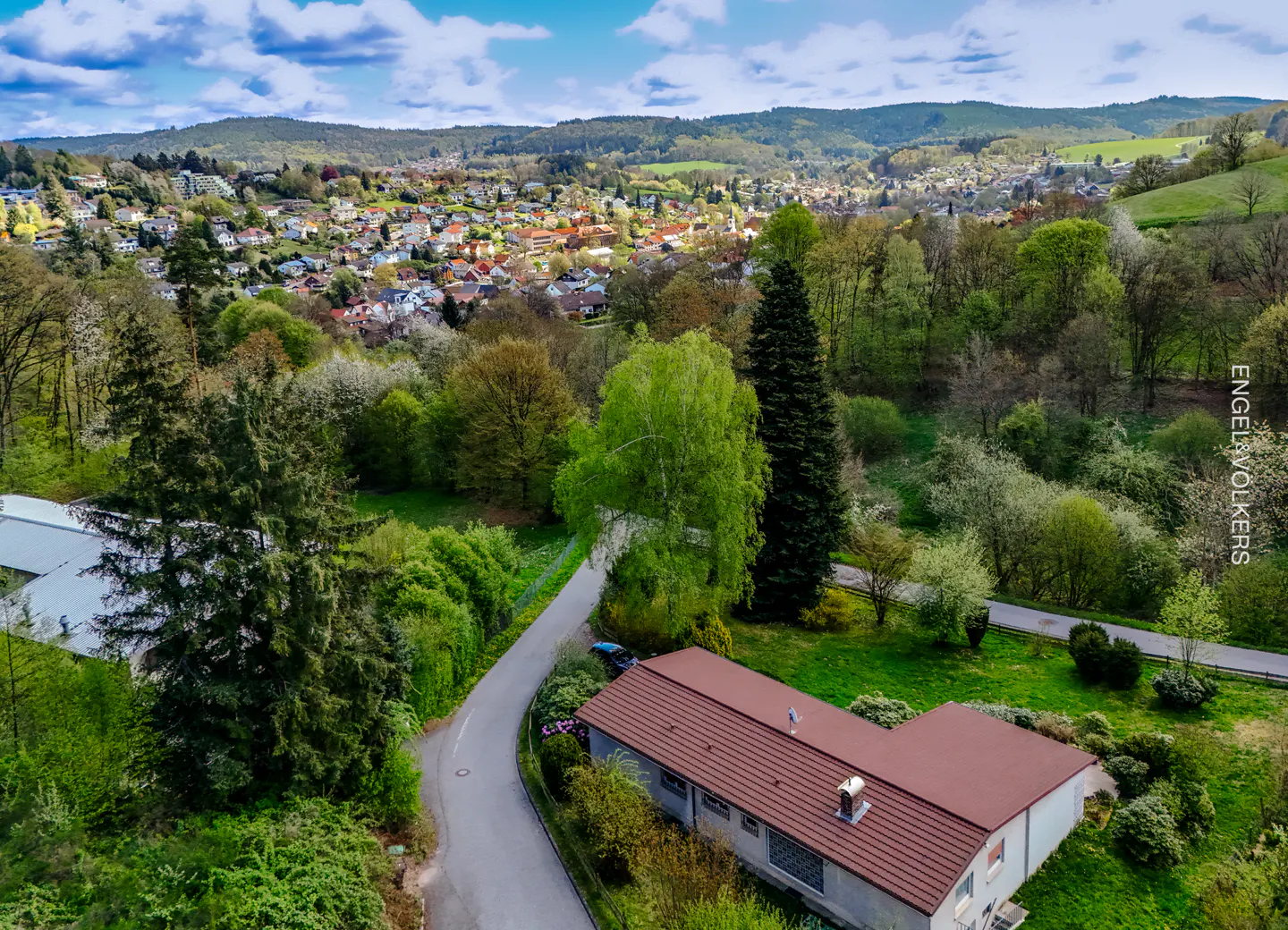 Aerial view of a house with a red roof, surrounded by green trees and a winding road, with a town in the background.