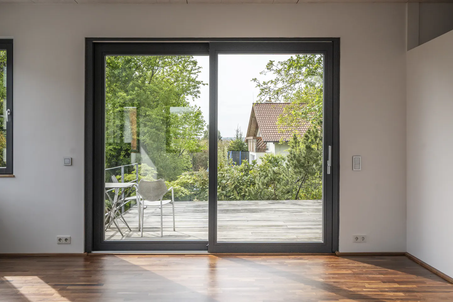 Interior view of a room with hardwood floors and a sliding glass door. Outside, a patio with chairs and a view of a house with a red tile roof.