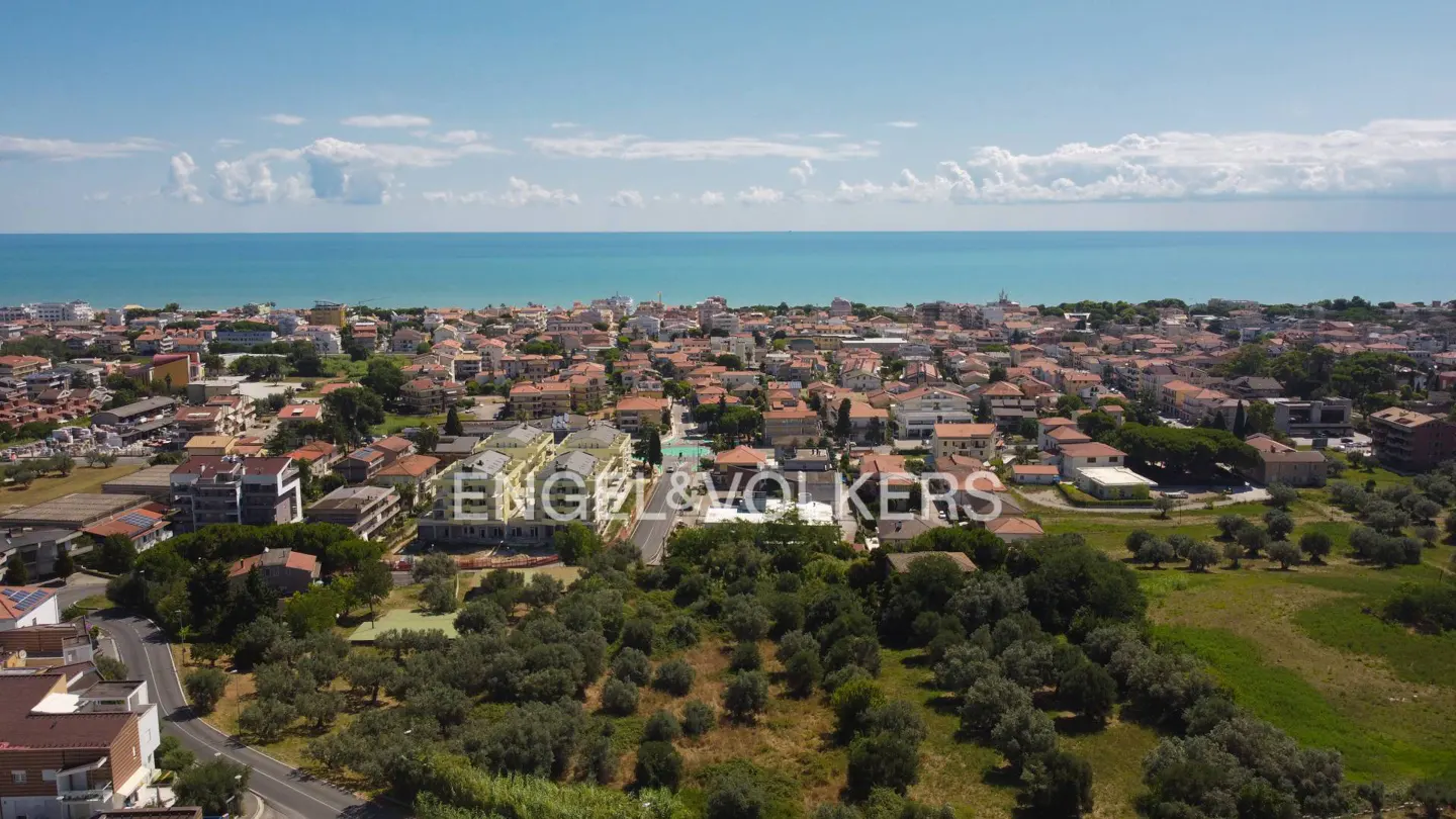 Aerial view of a coastal town with red-roofed buildings, green trees, and the blue sea under a partly cloudy sky. Engel & Volkers logo visible.