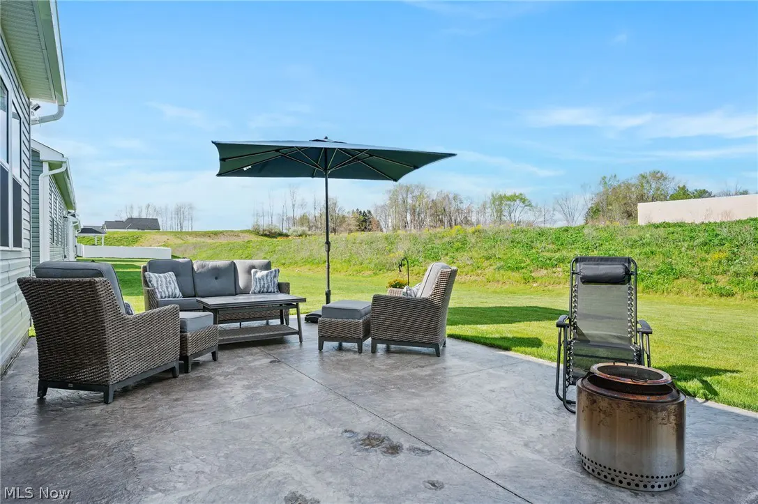 Outdoor patio with gray wicker furniture, umbrella, and fire pit on a concrete patio with a grassy lawn and blue sky.