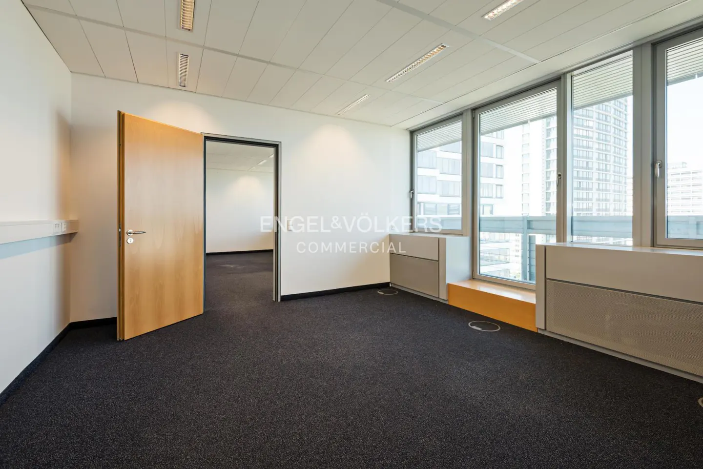 Empty office space with black carpet, white walls, and a light wood door. Large windows offer a city view.