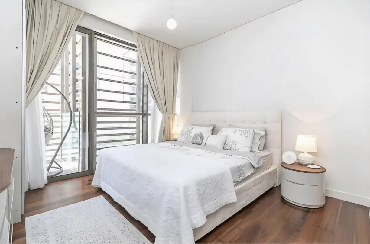Bright bedroom with white walls, wood floors, and a large window with curtains. A white bed with decorative pillows and a nightstand with a lamp are visible.