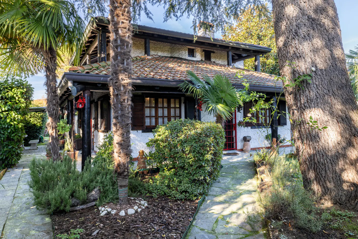 Exterior view of a two-story house with a red tile roof, white walls, and dark brown trim surrounded by trees and greenery.