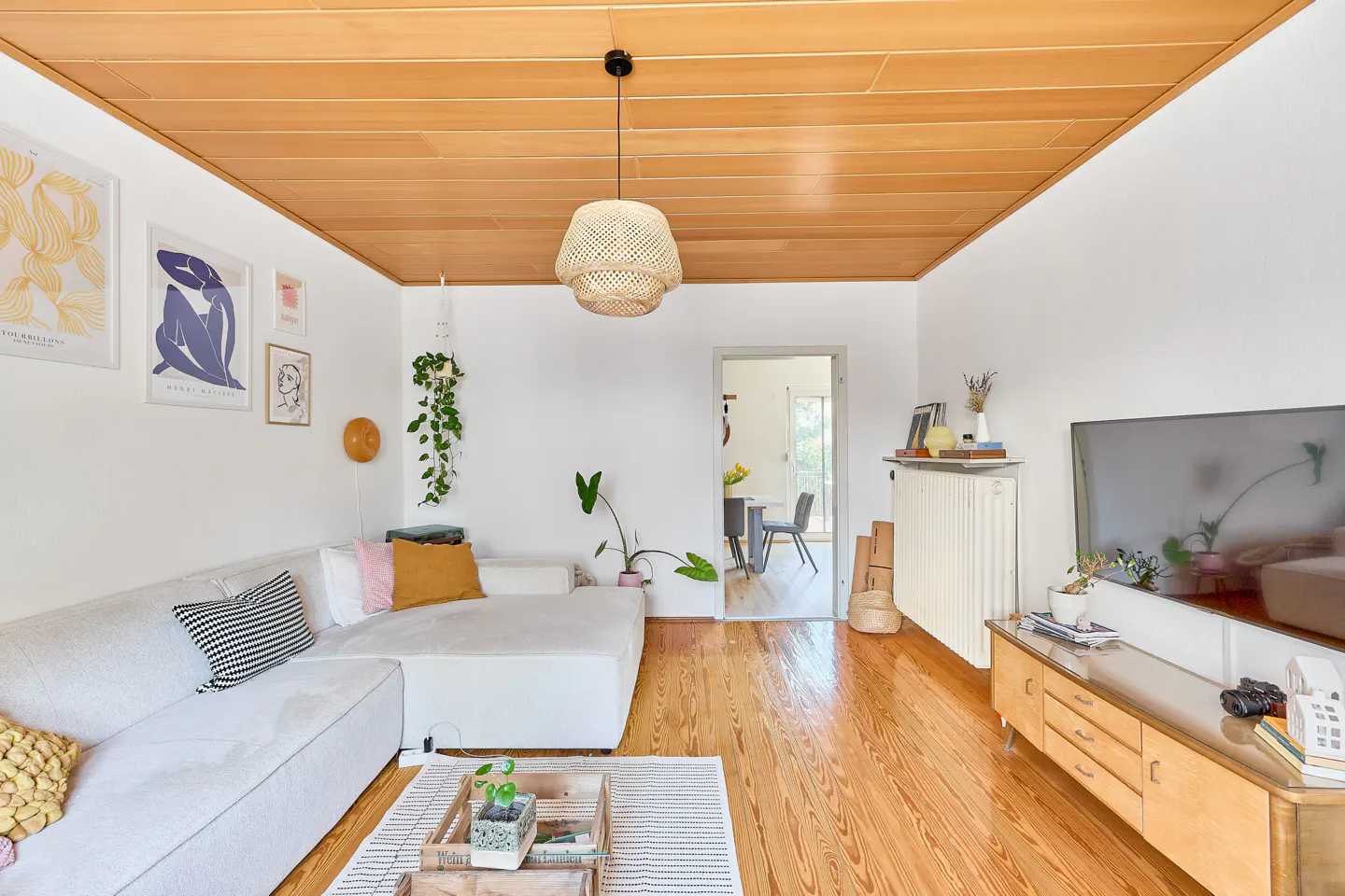Bright living room with wood floors, white walls, and a wood-paneled ceiling. A white sofa faces a TV on a mid-century modern console.
