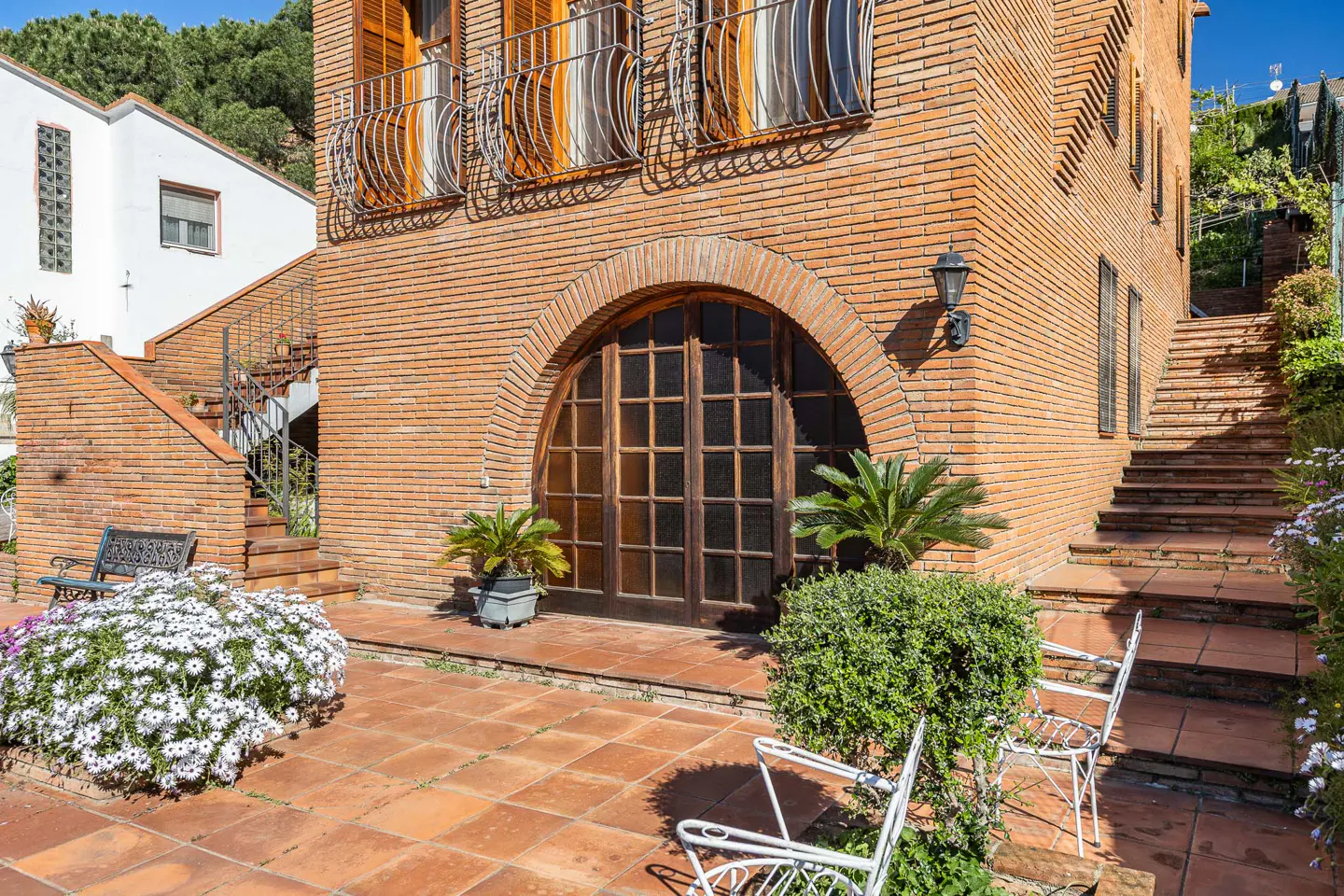 Exterior view of a two-story brick house with an arched wooden door, balconies, and a tiled patio with white chairs and flowers.