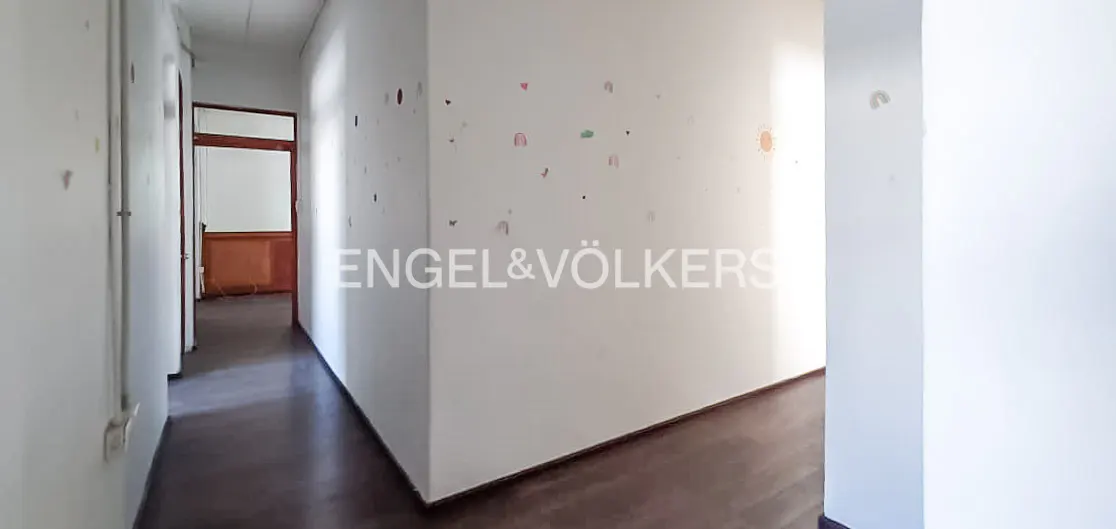 A bright hallway with white walls, dark wood floors, and an open doorway. The Engel & Völkers logo is visible on the wall.