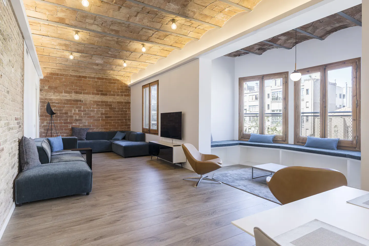 Bright living room with brick wall, wood floors, and large windows. A blue sectional sofa and tan chairs furnish the space.