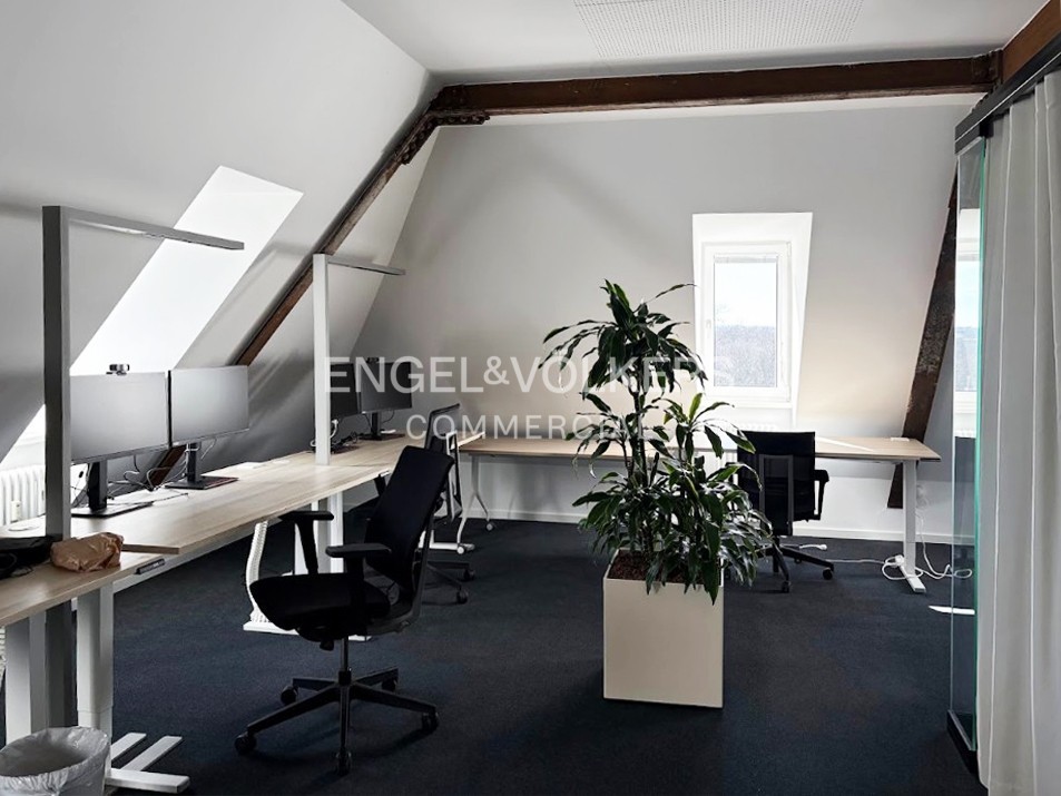 Modern attic office with slanted ceilings, wooden beams, light wood desks, black chairs, and a large potted plant.