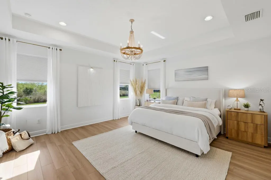 Bright, airy bedroom with white walls, wood floors, and a large bed. A chandelier hangs above a neutral rug. Windows offer a view of greenery.