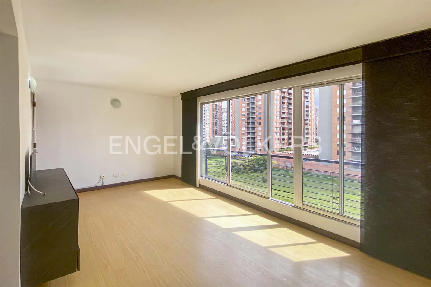Empty room with light wood floors, white walls, and a large window overlooking a green area and tall apartment buildings.