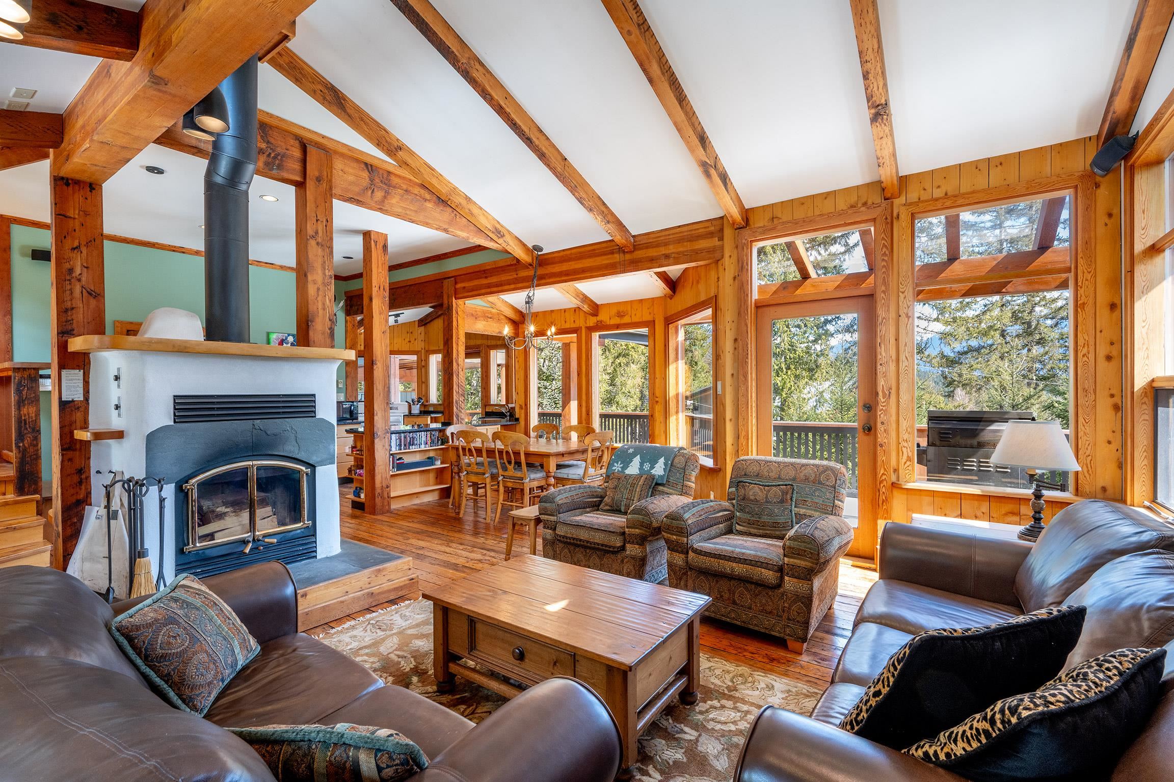 Living room with wood beams, fireplace, and leather sofas. Large windows overlook trees.