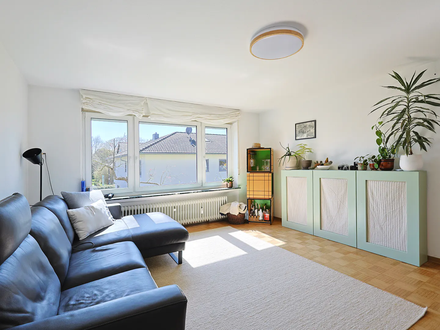 Bright living room with a black leather sectional sofa, white rug, and light green cabinets with plants on top. A large window lets in natural light.