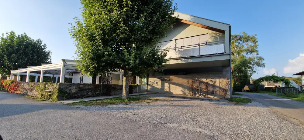 Modern two-story house with a balcony, a white pergola, and a large tree in the front yard. The house is beige and gray.