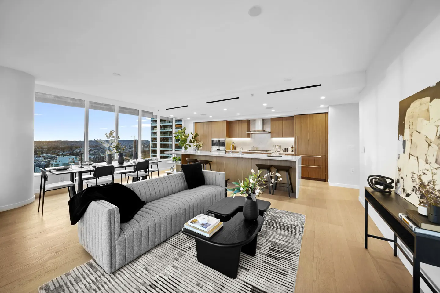 Bright, modern condo interior with a gray sofa, black coffee tables, and a dining area by a large window overlooking the city. The kitchen features wood cabinets and a white island.