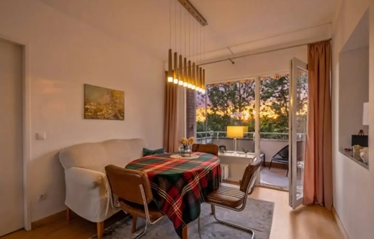 Dining area with a plaid-covered table, two leather chairs, a white sofa, and a balcony view at sunset.