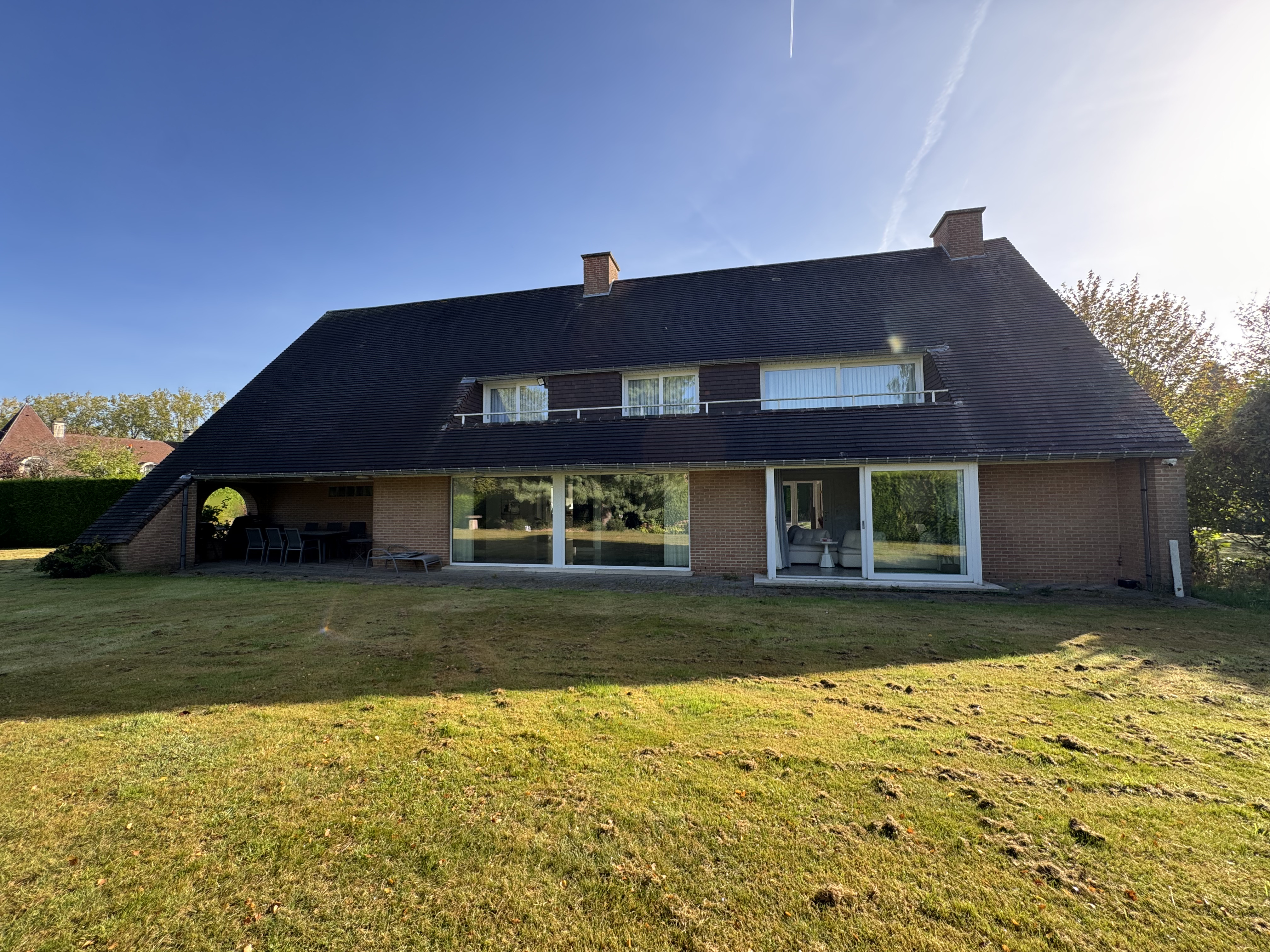 Large brick house with a steep dark roof, sliding glass doors, and a covered patio overlooking a sunny green lawn. [AI-generated text]