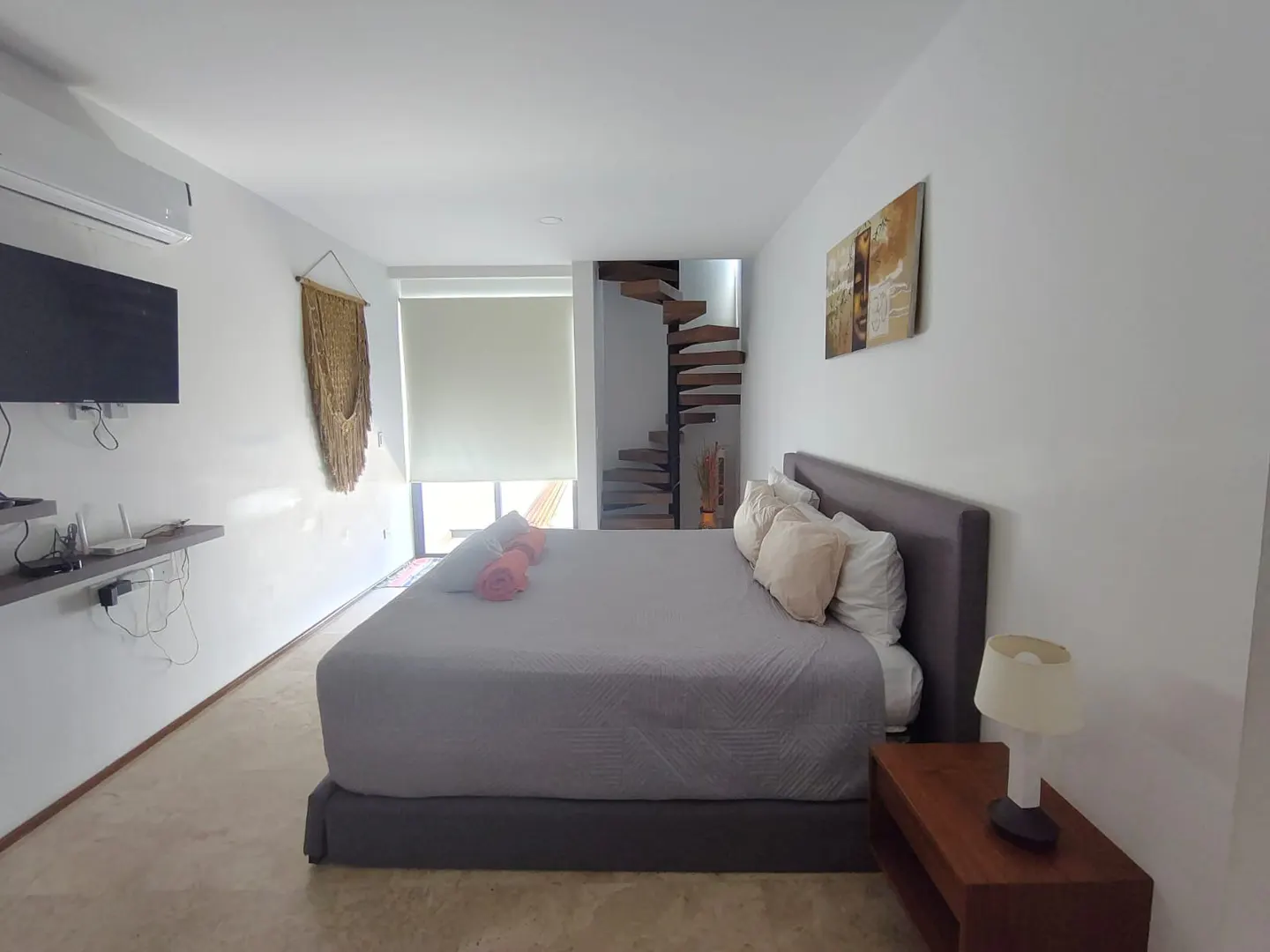 Bedroom with a gray bed, white walls, and a spiral staircase. A TV and macrame wall hanging are on the left wall. A painting hangs on the right wall.