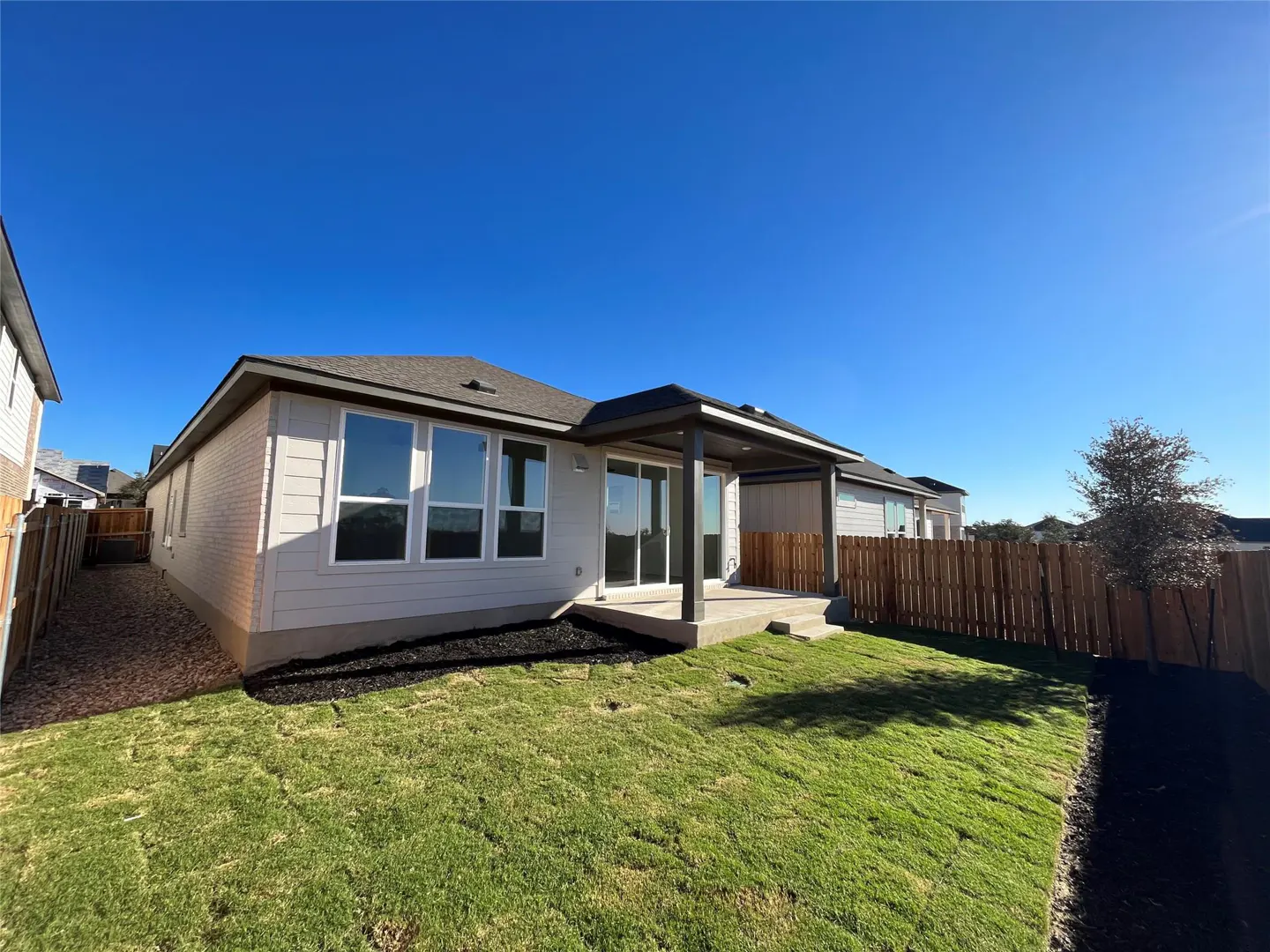 Backyard view of a single-story house with a covered patio, green lawn, and wooden fence under a clear blue sky.