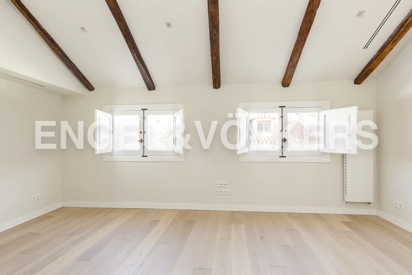 Bright, empty room with light wood floors, white walls, and exposed beams. Two windows with white shutters flank the Engel & Völkers logo.