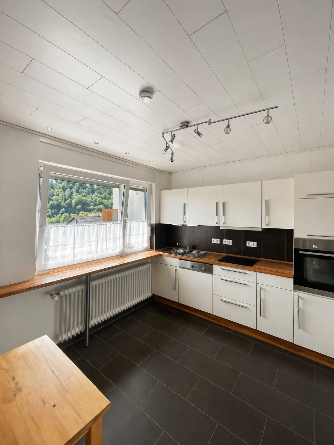 Bright kitchen with white cabinets, black backsplash, and wood countertop. A window with sheer curtains overlooks greenery. Dark tile floor and wooden table.
