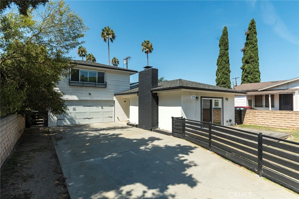 White two-story house with a black brick chimney, concrete driveway, and black horizontal fence under a clear blue sky. [AI-generated text]