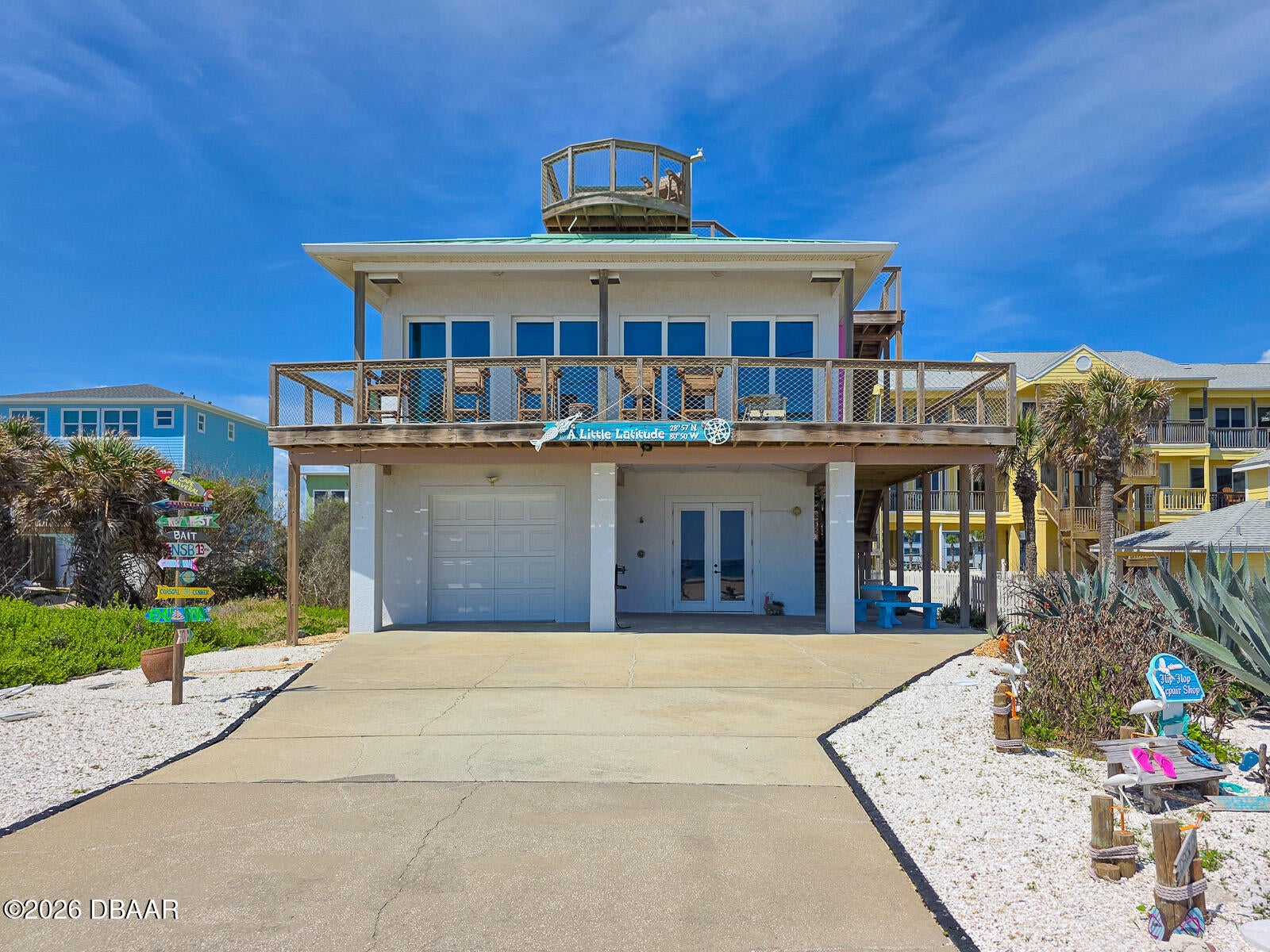 Two-story white beach house on stilts with a rooftop deck, large balcony, and concrete driveway under a bright blue sky. [AI-generated text]