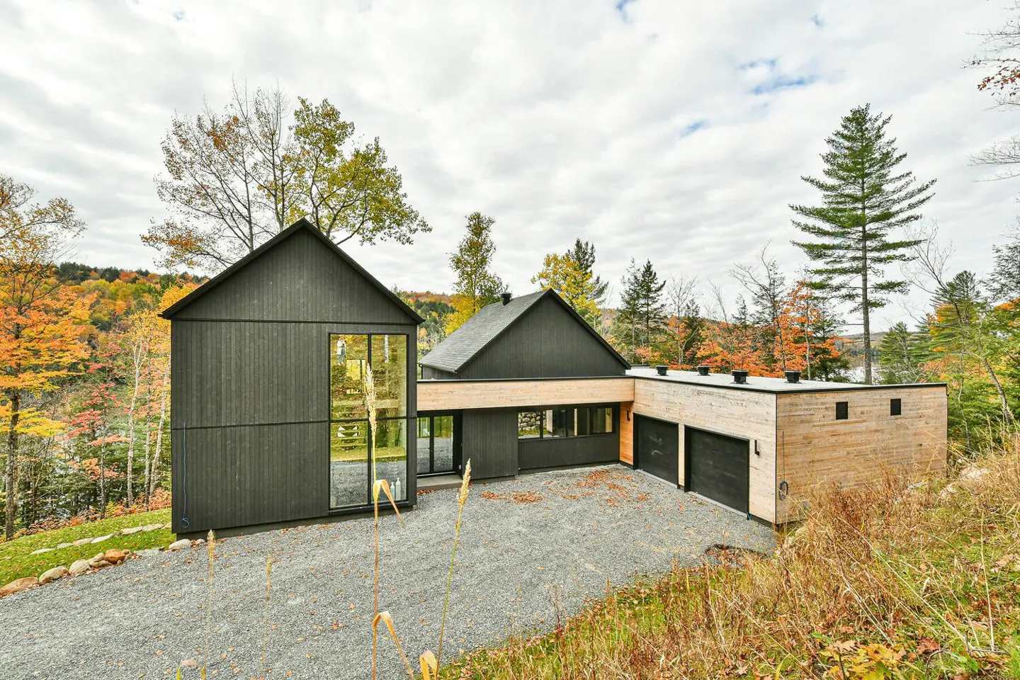 Modern black and wood home with a two-car garage on a gravel driveway, surrounded by fall foliage.