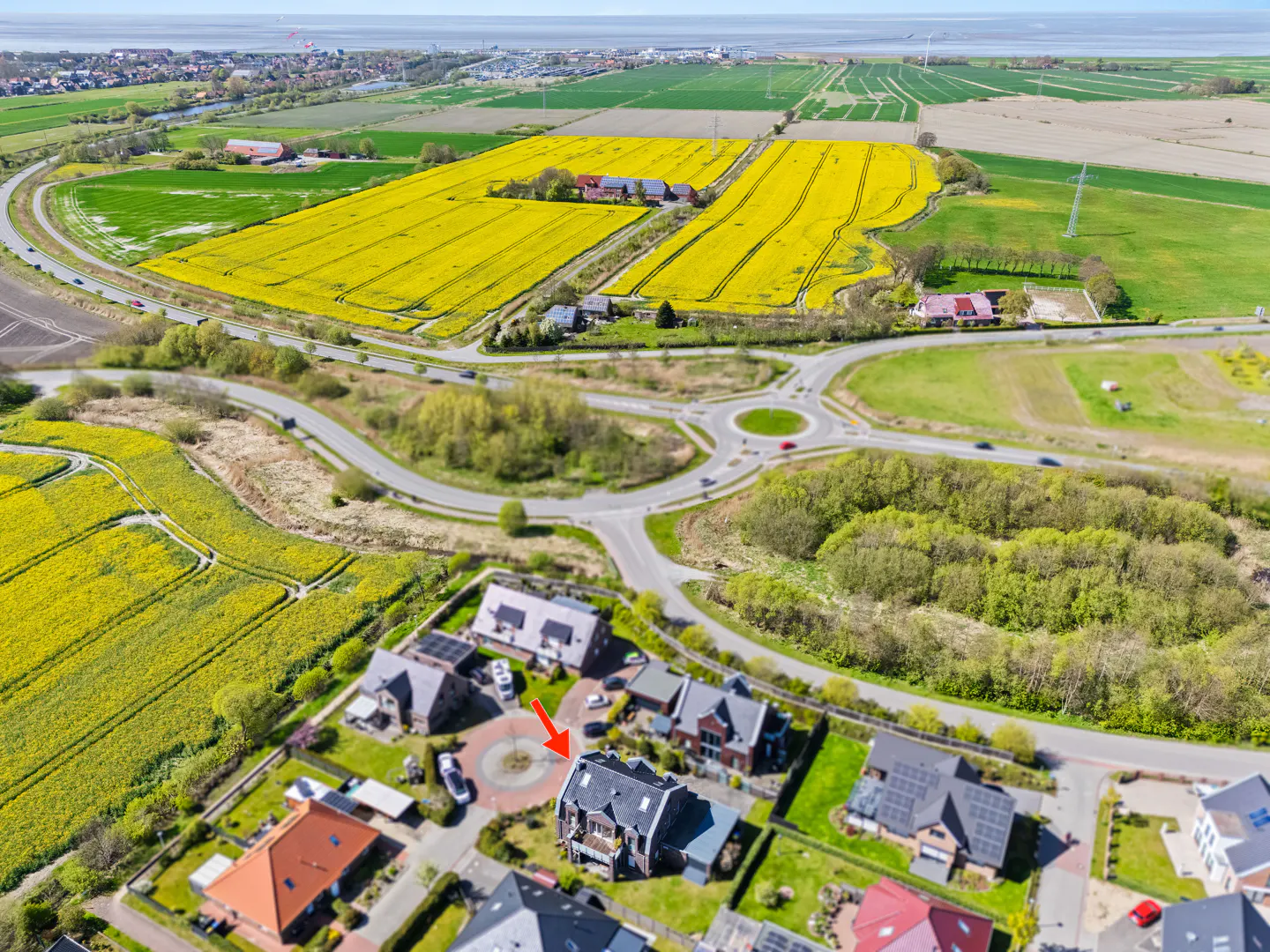 Aerial view of a dark-roofed house with a circular driveway, surrounded by green lawns and other houses, near yellow fields and a roundabout.