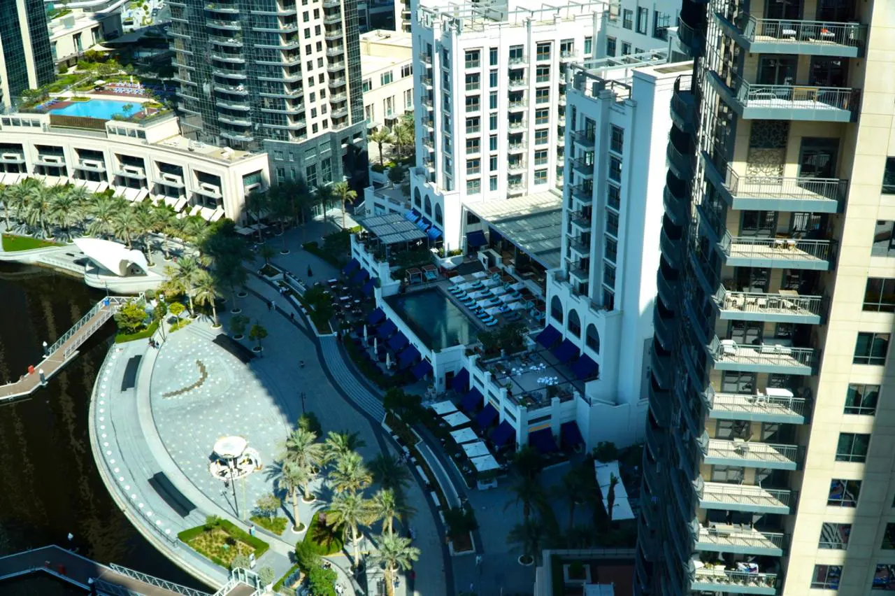 Aerial view of a luxury hotel with a pool, blue awnings, and palm trees, surrounded by modern buildings in a city.