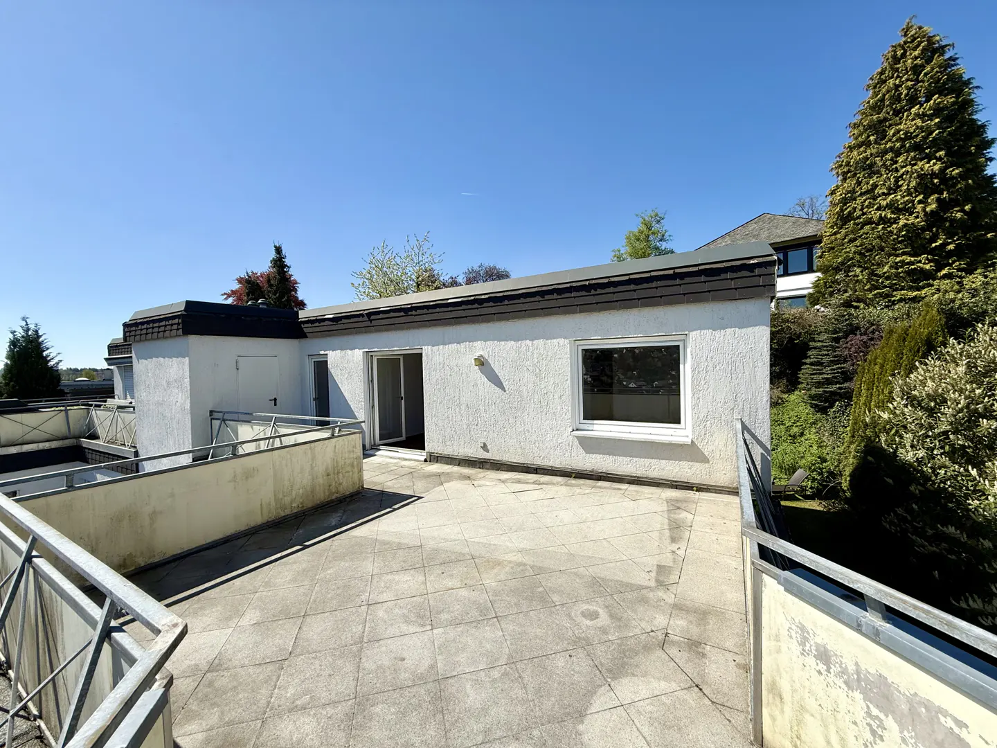 Exterior view of a white building with a patio, metal railings, and a blue sky. Trees and greenery are visible in the background.