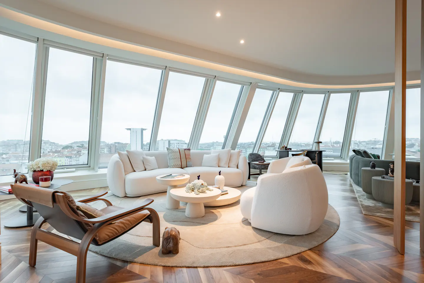 Bright living room with curved windows, a white sofa, and a round rug on a herringbone wood floor. A brown leather chair sits in the foreground.