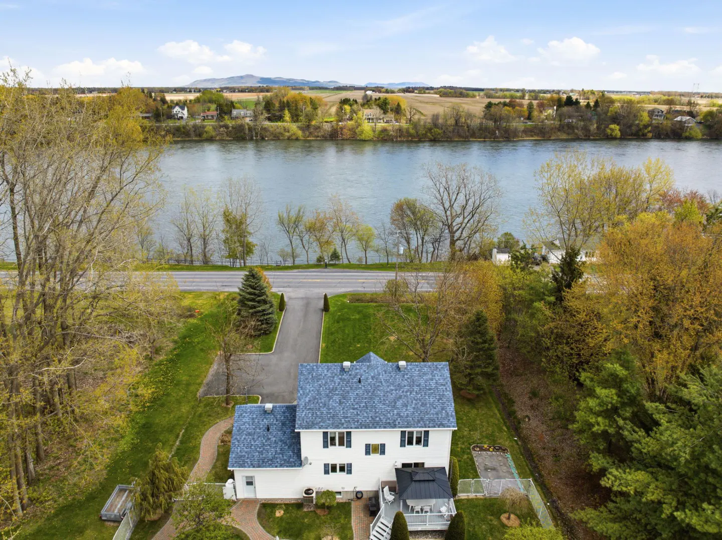 Aerial view of a white two-story house with a blue roof, near a river and green trees under a blue sky.