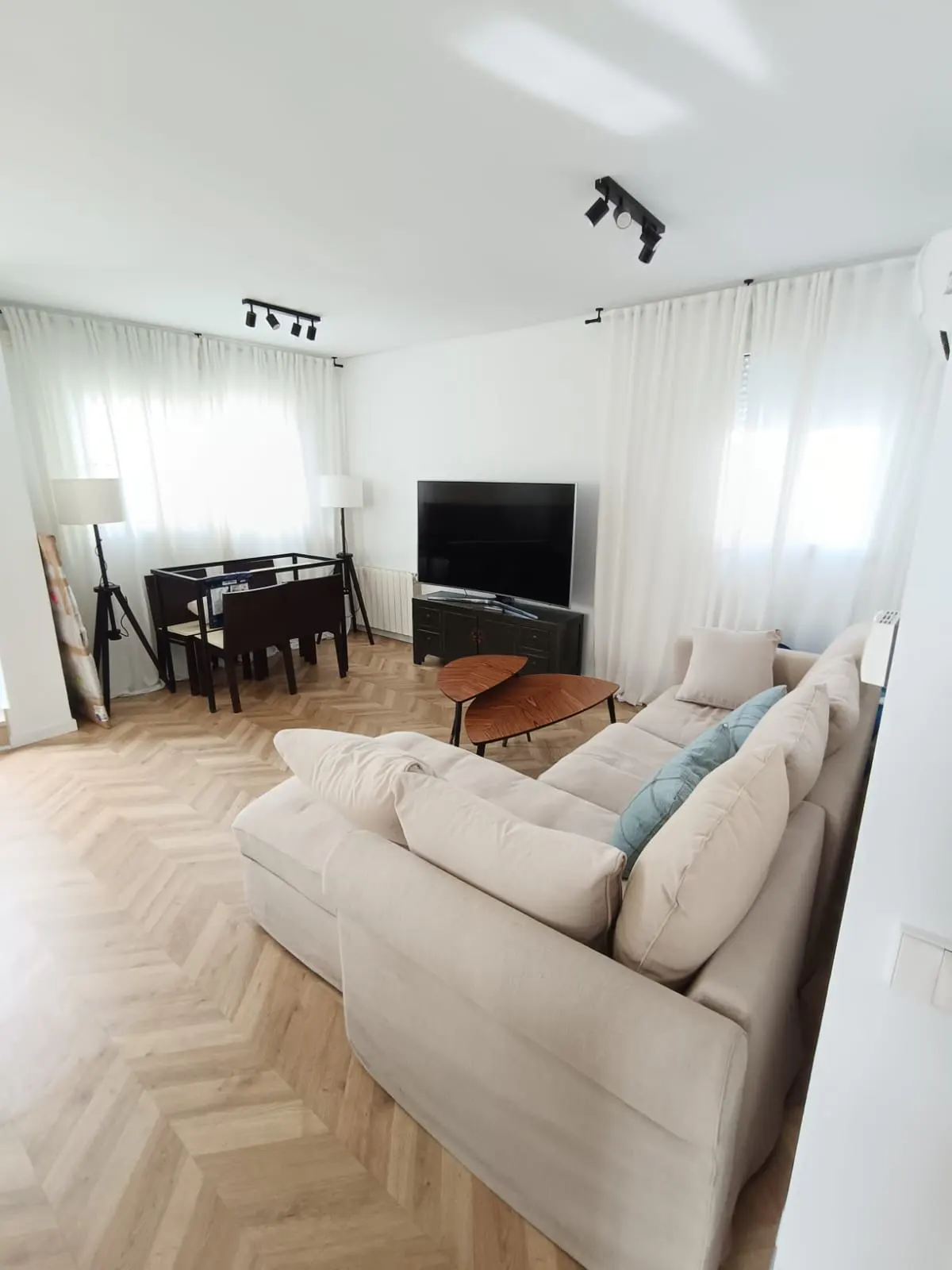 Bright living room with a beige sectional sofa, wooden coffee table, TV, and foosball table on a herringbone wood floor. White walls and curtains.