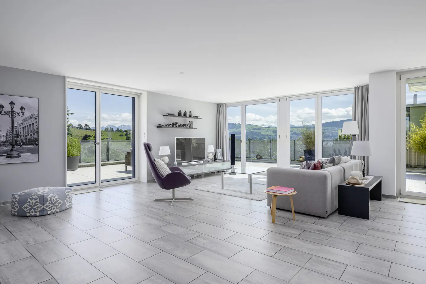 Bright, modern living room with gray tile floors, a purple chair, and a gray sofa. Large windows offer a view of green hills.