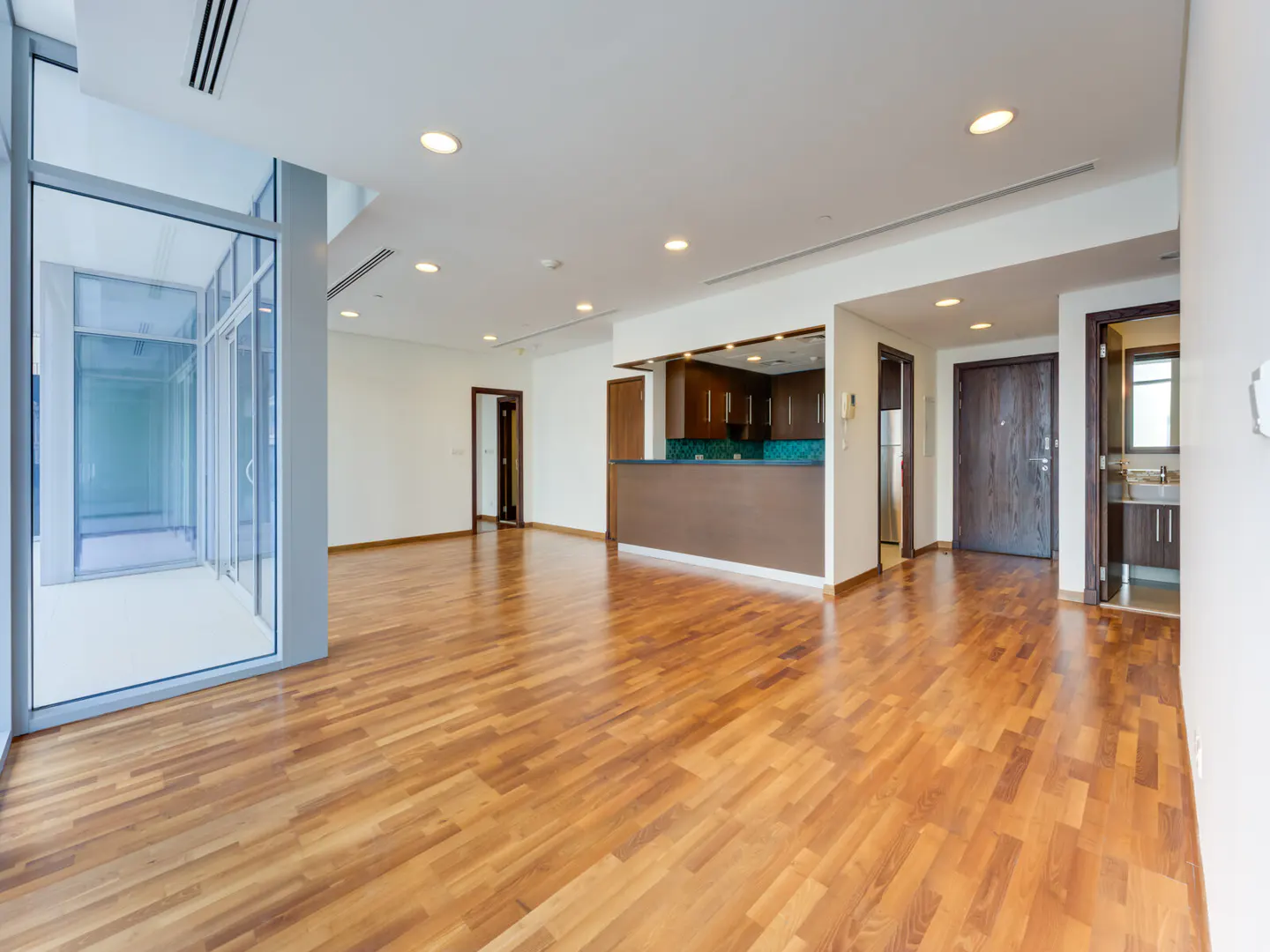 Bright, empty apartment with hardwood floors, white walls, and recessed lighting. A kitchen area with brown cabinets and a teal backsplash is visible.
