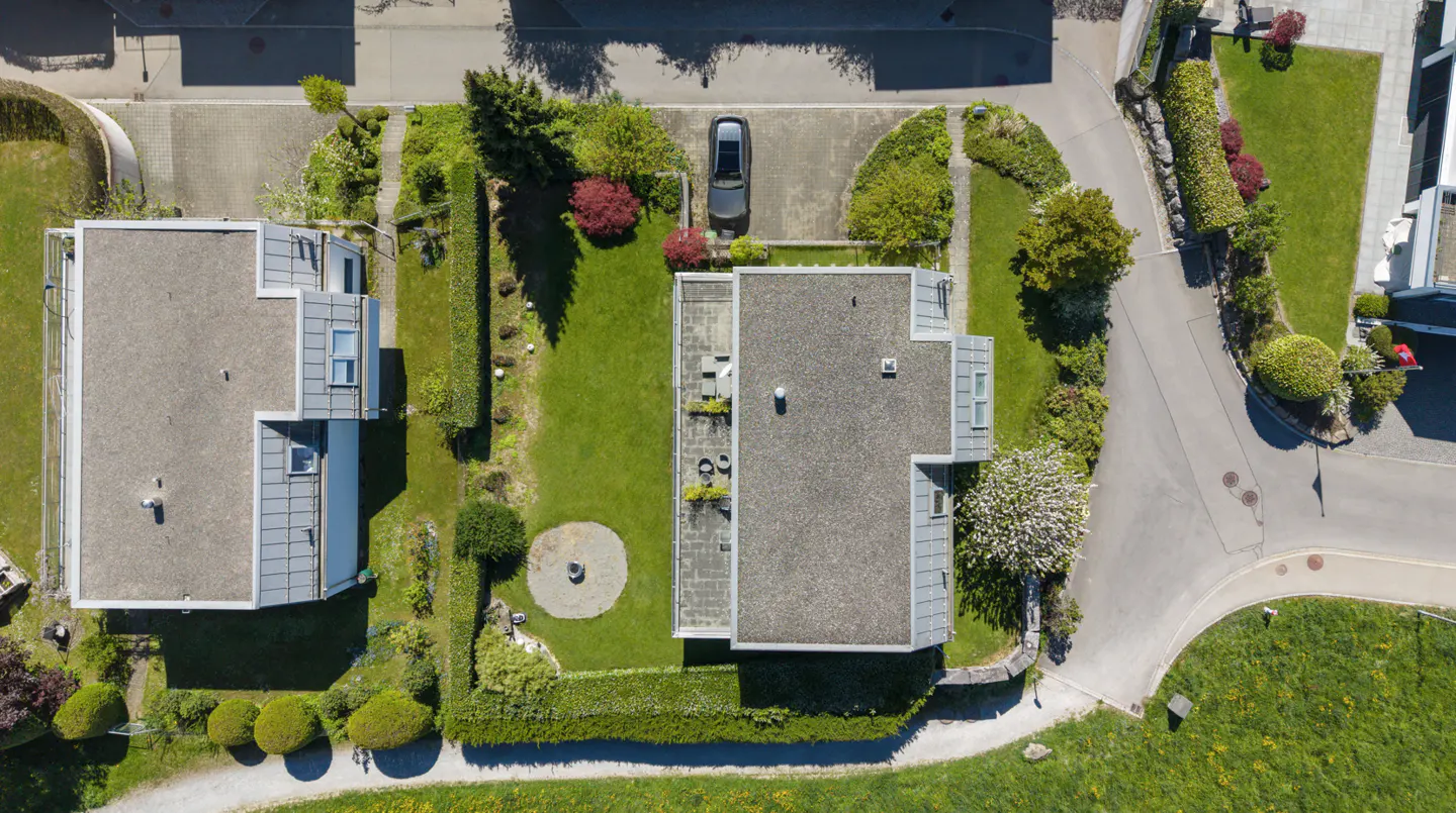 Aerial view of two modern houses with gray roofs, green lawns, trees, and a parked car.