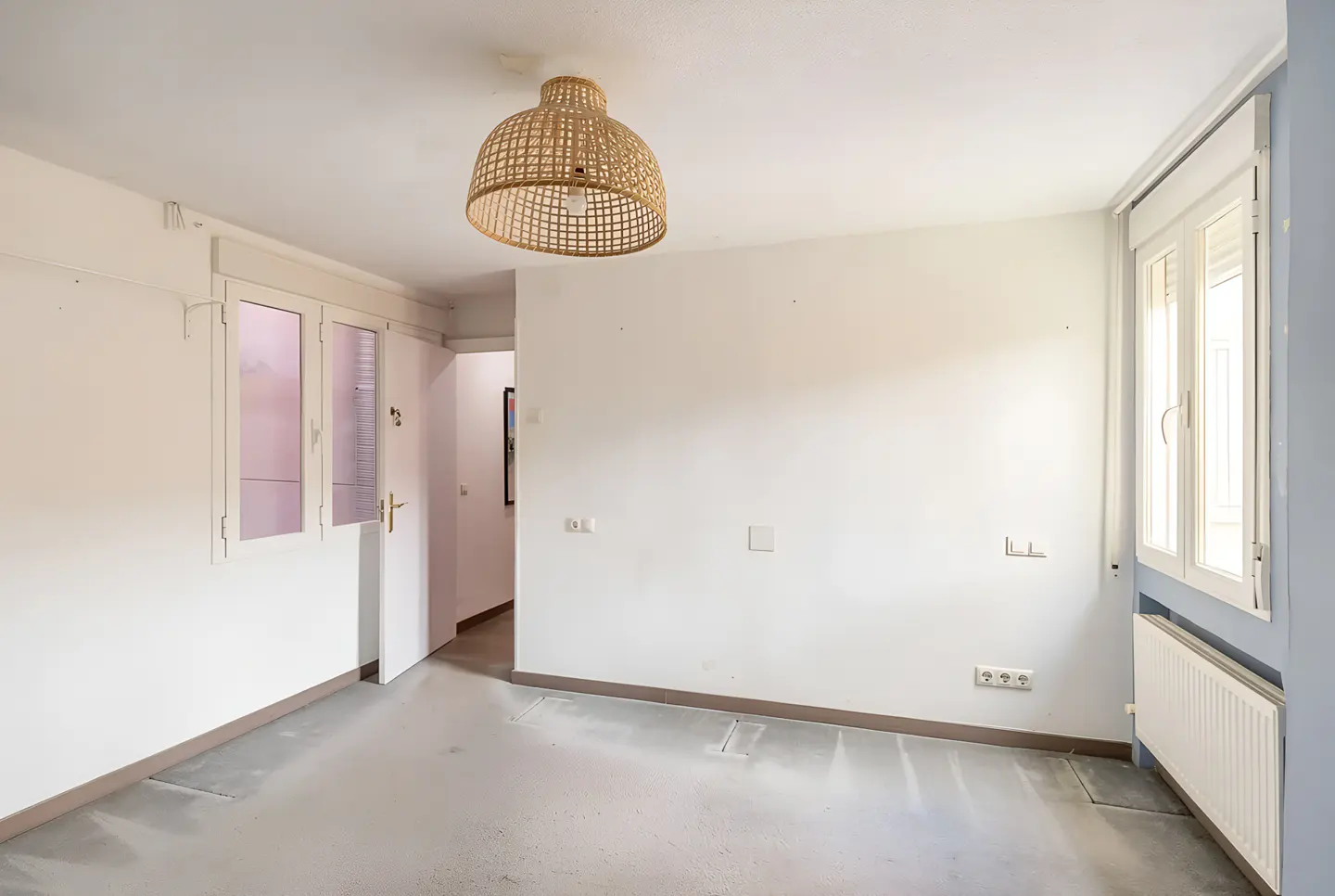 A bright, empty room with white walls, a window, and a woven pendant light.