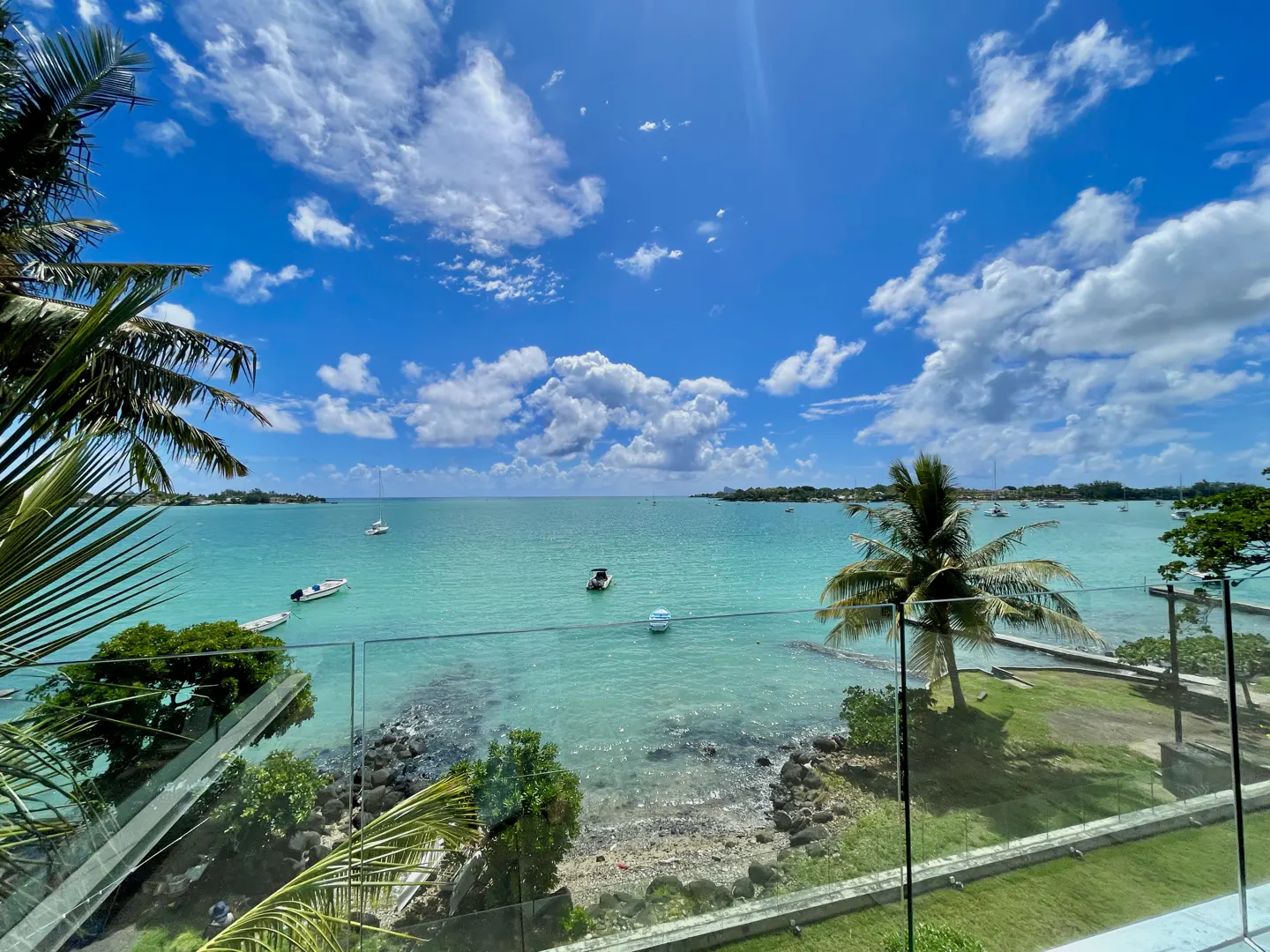 Turquoise ocean view from a balcony with palm trees, boats, and a bright blue sky with white clouds.