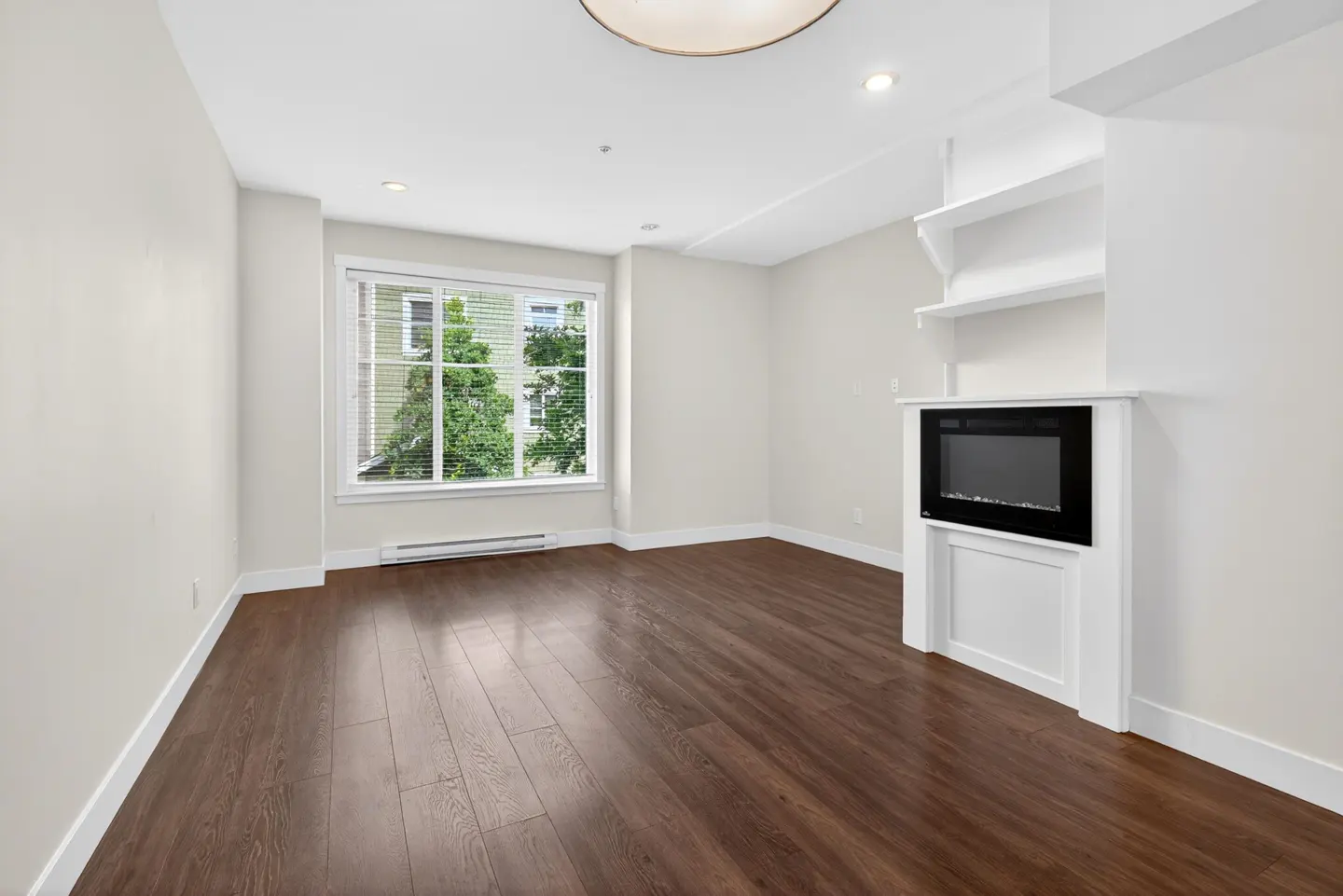 Bright living room with dark wood floors, white walls, a fireplace with built-in shelving, and a large window with greenery.