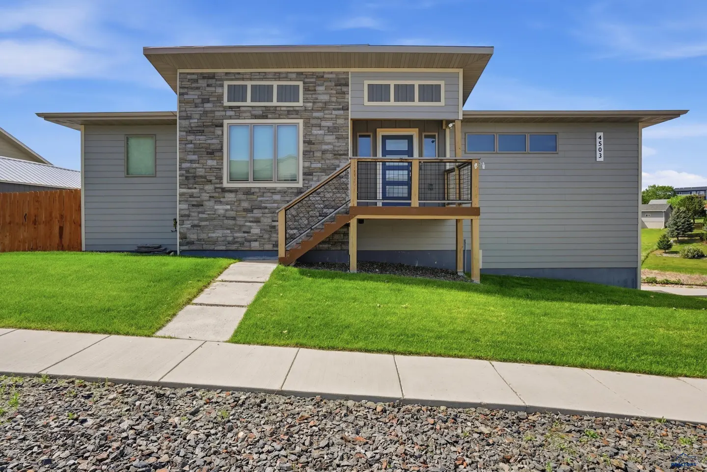 Modern gray home with stone facade, elevated front porch, and blue door, set on a green lawn under a clear blue sky.
