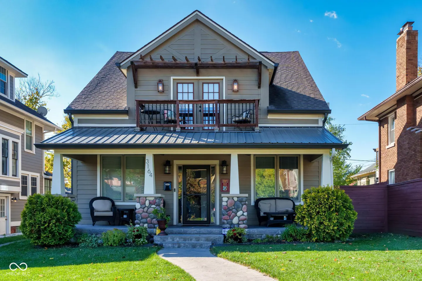 Two-story house with a balcony, stone porch columns, and green lawn under a blue sky.