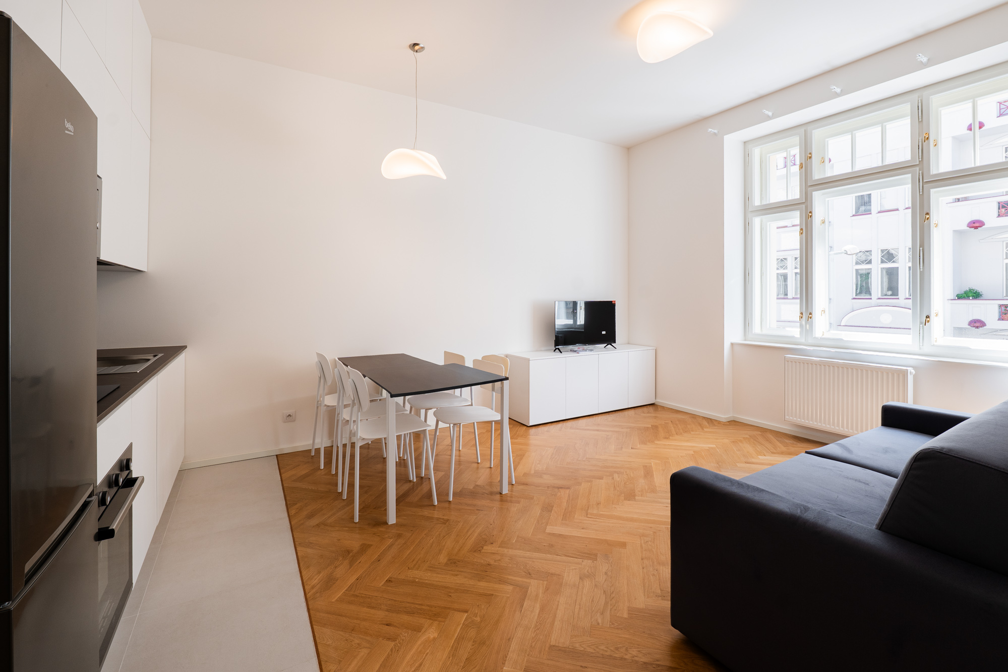 Modern studio apartment with a black sofa, dining table, white kitchen cabinets, and herringbone wood floors.