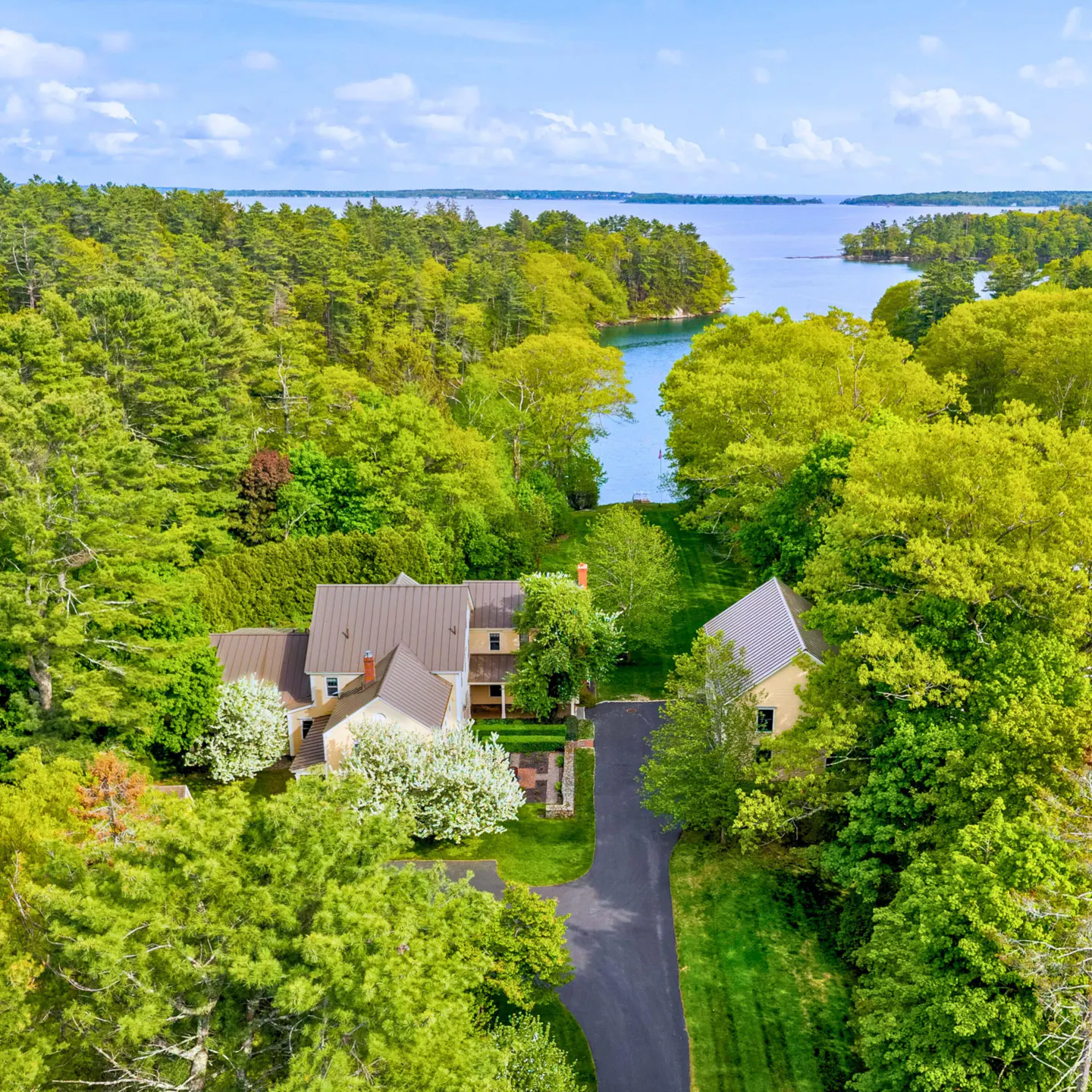Aerial view of a tan house with a brown roof, surrounded by green trees and a blue body of water.